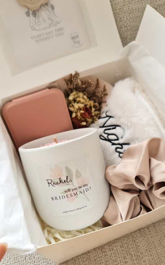 A gift set for bridesmaids including a customised candle, a proposal card, a scrunchie, a foldable mirror, and an eye cover, all displayed in an open gift box.