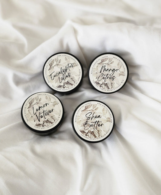 Mini Candles Discovery Set, Black Tins, Silk Ivory Candles, Four small containers with black lids are arranged on a white cloth background. Each lid has a decorative label illustrating different scents: Eucalyptus Lilies, Mango Citrus, Lemon Vetiver, and Shea Butter. The labels feature botanical designs in soft colors.