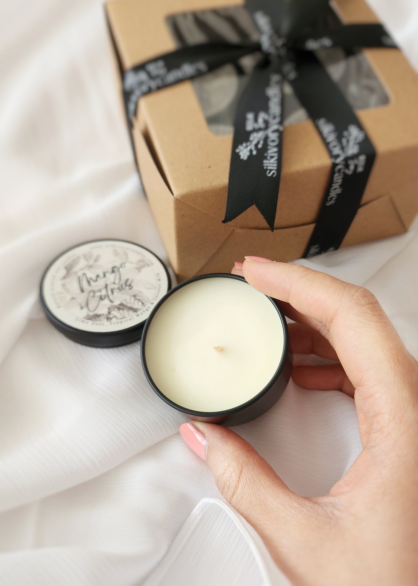 A hand holds an open tin candle with light beige wax, perfect for travel size convenience. The lid beside the candle reads "Luxury Candles." In the background, a brown gift box tied with a black ribbon rests on white fabric, ideal for presenting these elegant candle samplers.